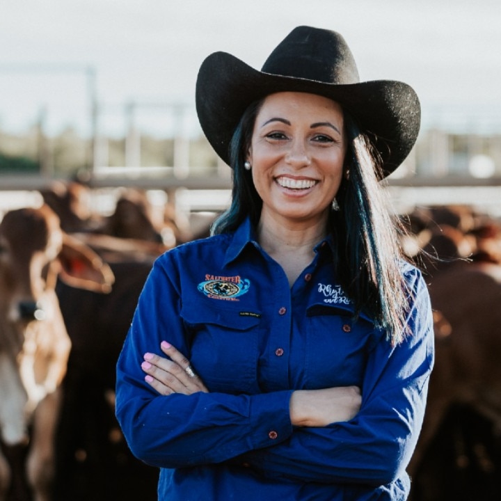 Cara Peek, wearing a cowgirl hat, stands in front a yard full of cattle