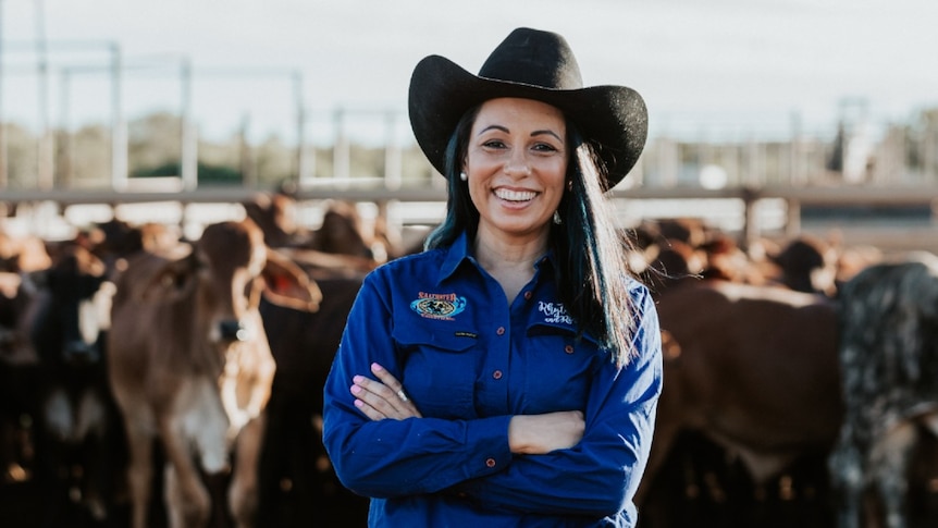 Cara Peek, wearing a cowgirl hat, stands in front a yard full of cattle
