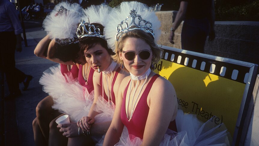 The Go-Go's wearing pearls, tutus and tiaras, sitting on a bench, one member smokes a cigarette
