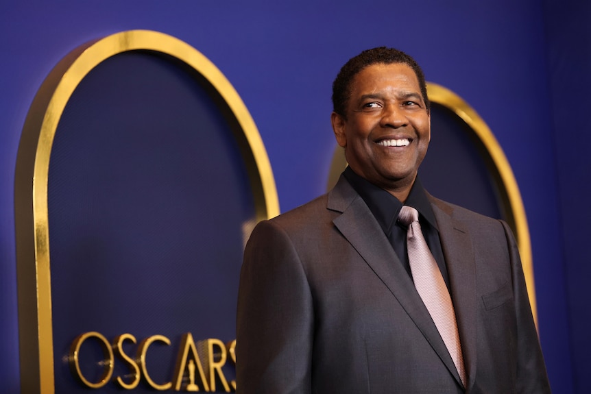 actor denzel washington smiles wearing a suit and tie in front of a blue and gold oscars branded background