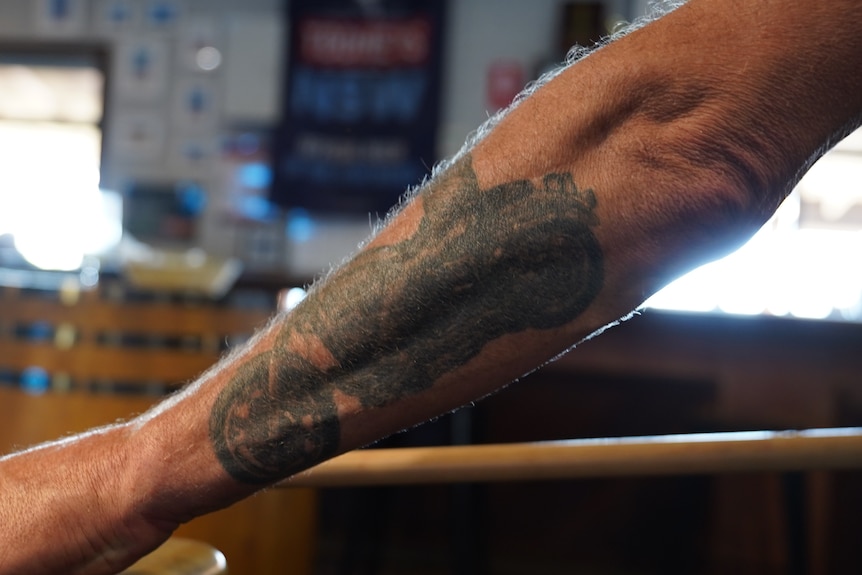 An oustretched arm over a pool table. There is a tattoo of a motorbike on the forearm.