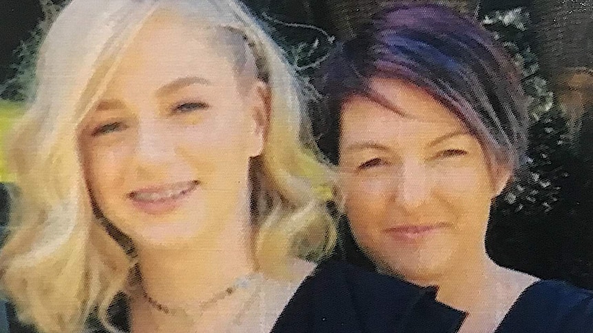 A blonde girl with shoulder length hair stands in front of a woman with dark short hair. Both smile at the camera.