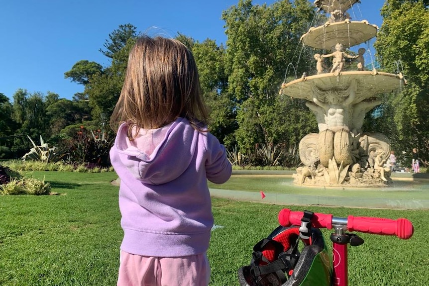 A small child with her back turned stands next to a scooter in a park.