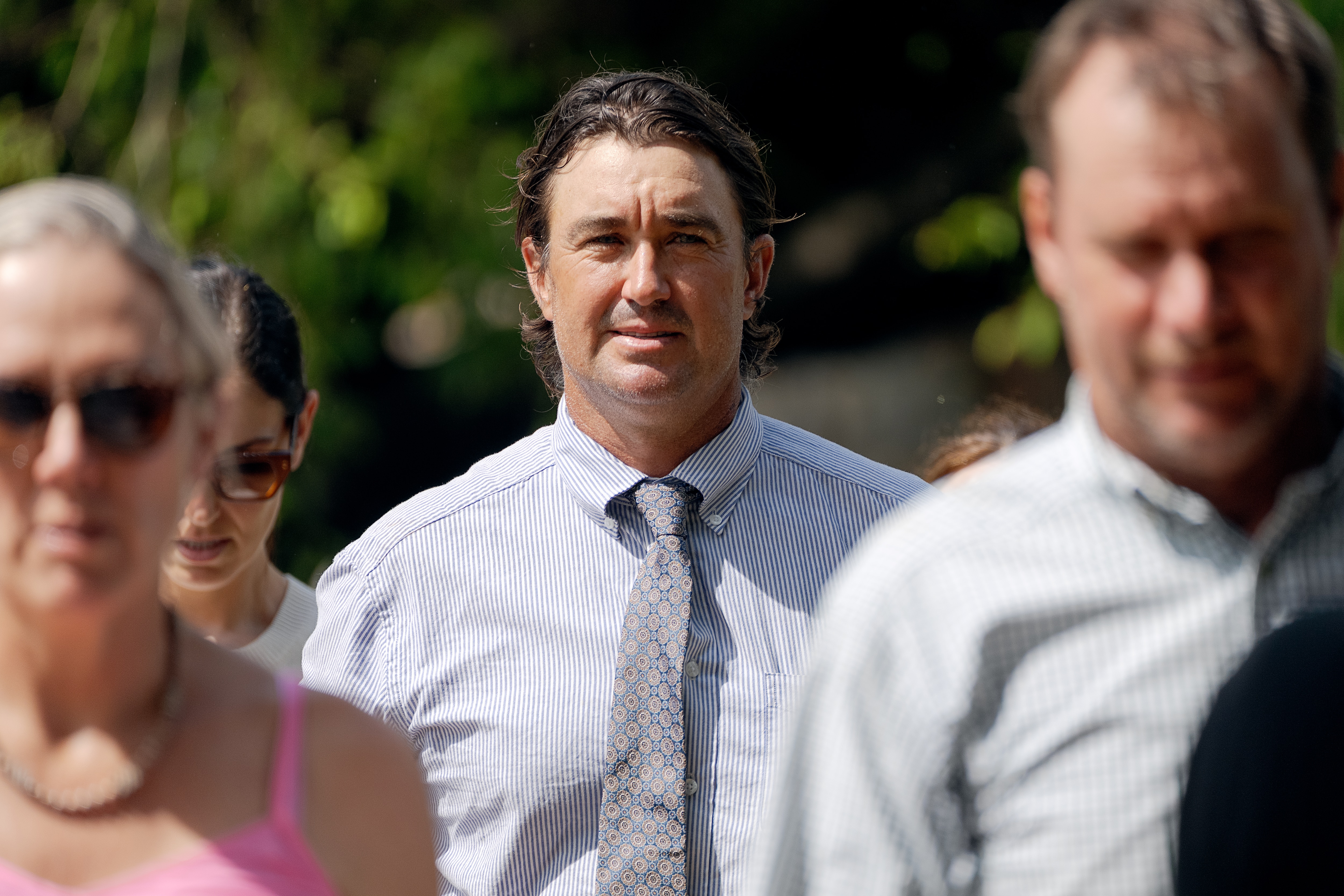 A white man in a button up shirt and gray tie, collar length brown hair, in focus as people walk all around him.