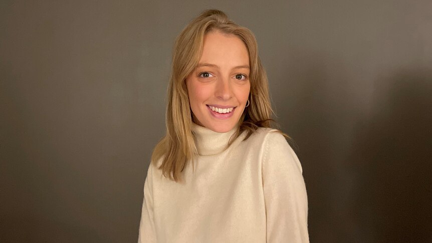 A woman with blonde hair and a cream jumper smiling against a brown backdrop.