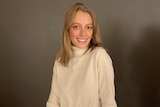 A woman with blonde hair and a cream jumper smiling against a brown backdrop.
