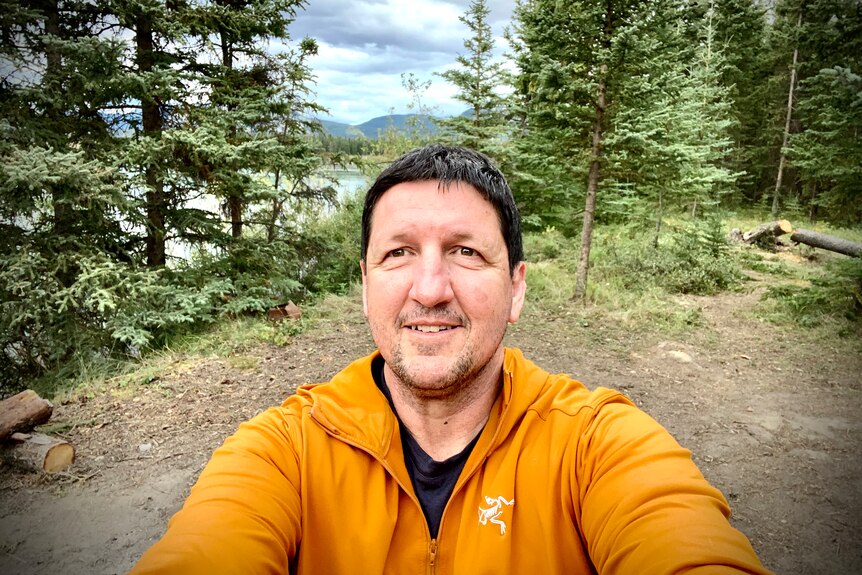 A man in a yellow puffer jacket takes a selfie with trees behind him.
