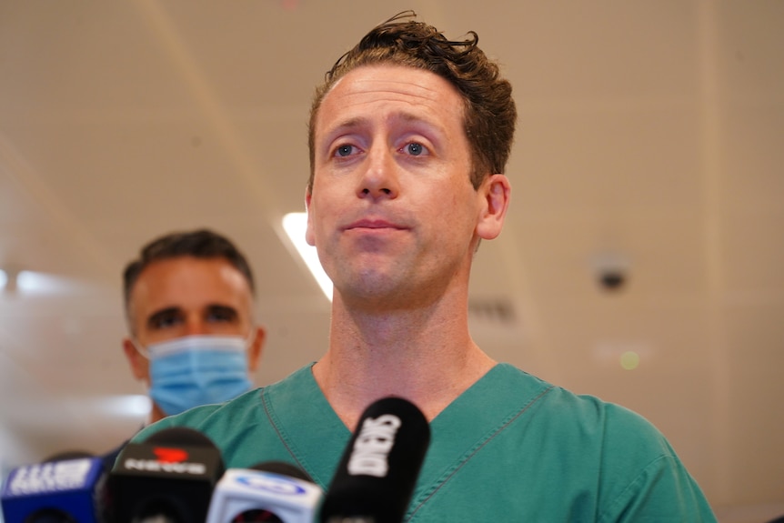 A man wearing green hospital scrubs stands in front of a microphone. Behind him is a man in a suit and face mask