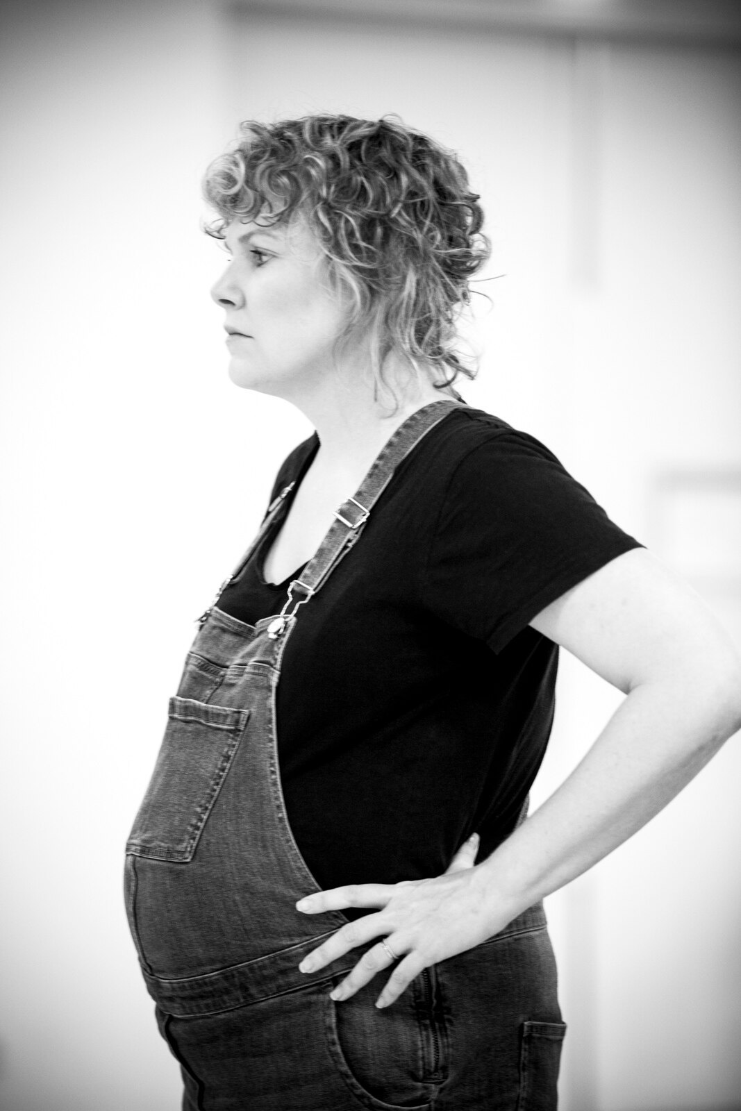 A B&W image of a pregnant, blonde, curly-haired woman, wearing a black shirt and overalls, her hands on her hips