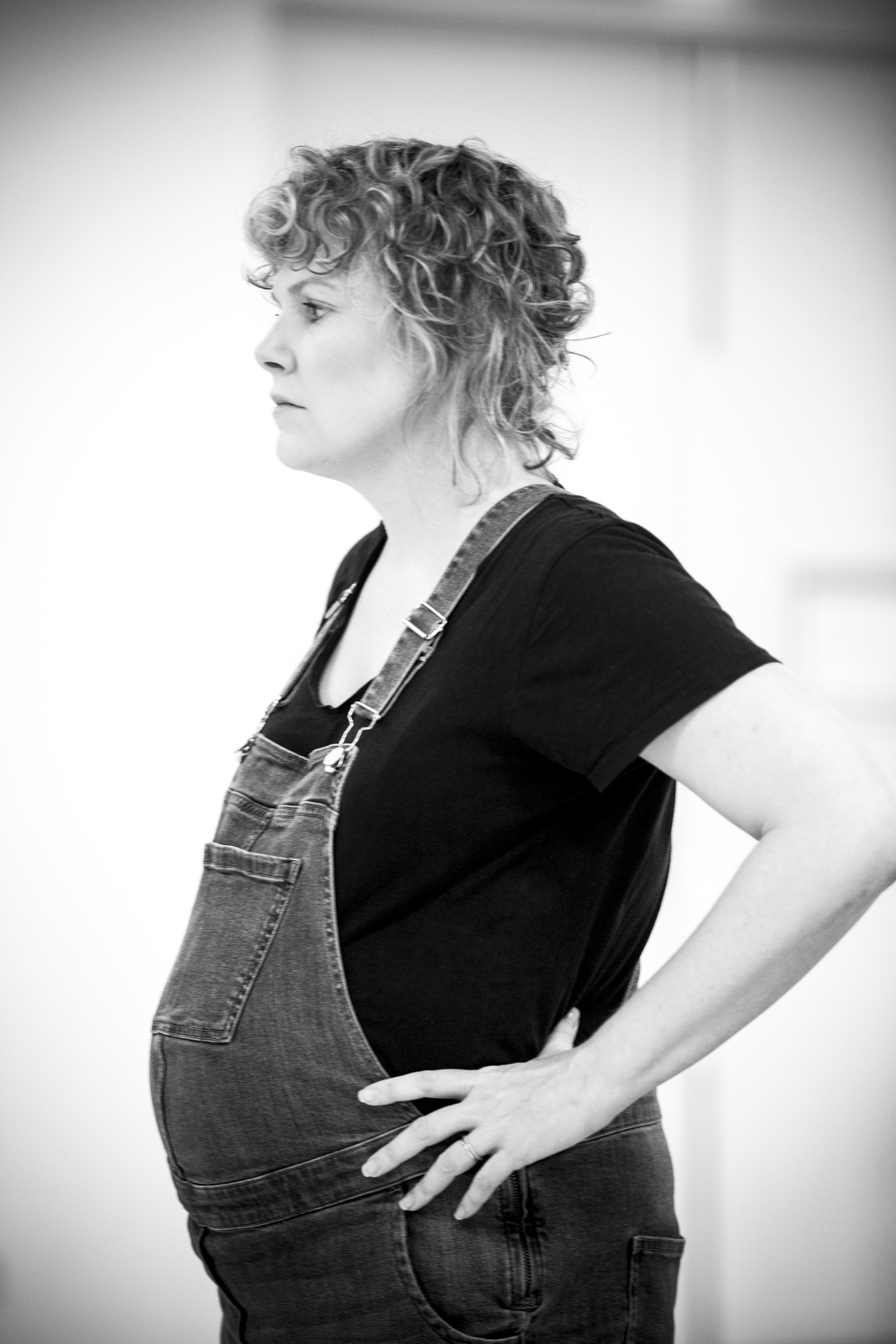 A B&W image of a pregnant, blonde, curly-haired woman, wearing a black shirt and overalls, her hands on her hips