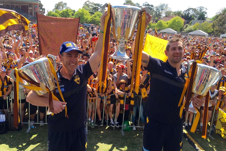 Alistair Clarkson and Luke Hodge hold up three premiership cups draped in brown and gold, with many fans in the background 