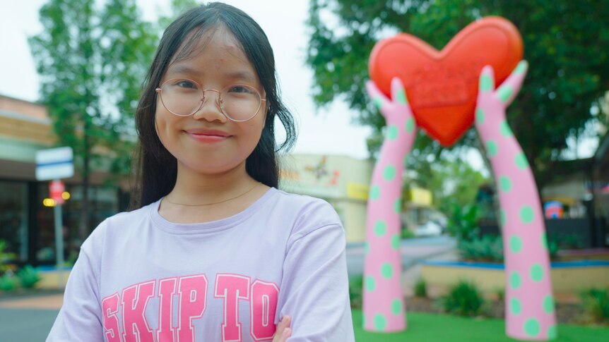 A young Vietnamese girl wearing round glasses smiles, wearing a purple t-shirt saying 'Skip to' 