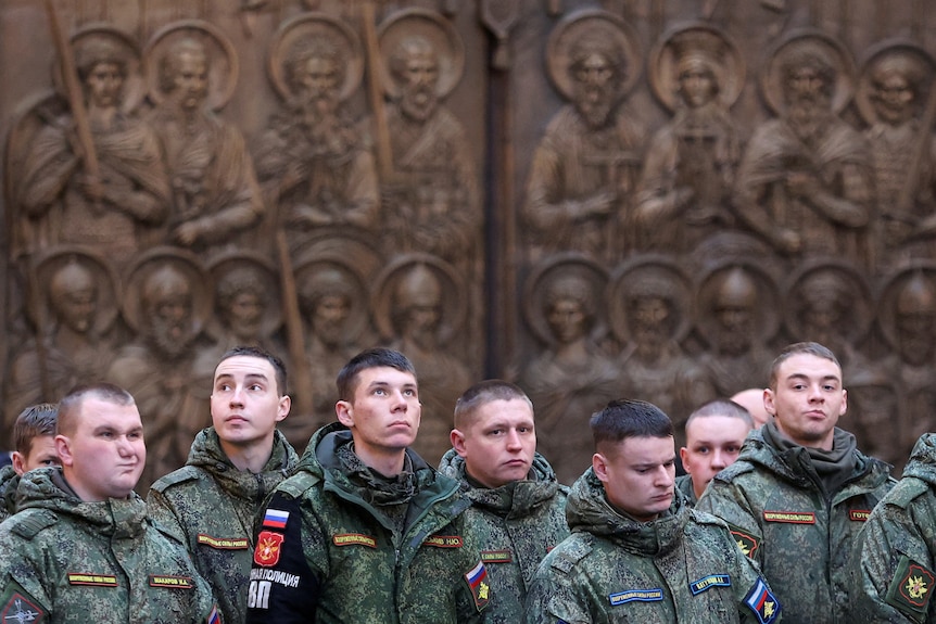 A group of young men in military uniform stand in front of an ornate, sculpted church wall.
