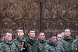 A group of young men in military uniform stand in front of an ornate, sculpted church wall.