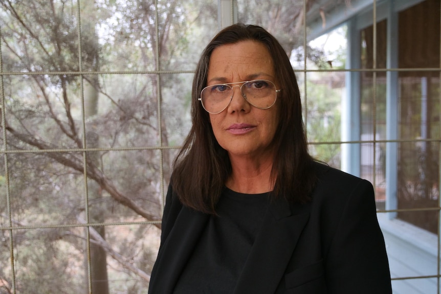 A serious woman with glasses and dark hair stands in an enclosed verandah, tree behind, blue paint on window frames.