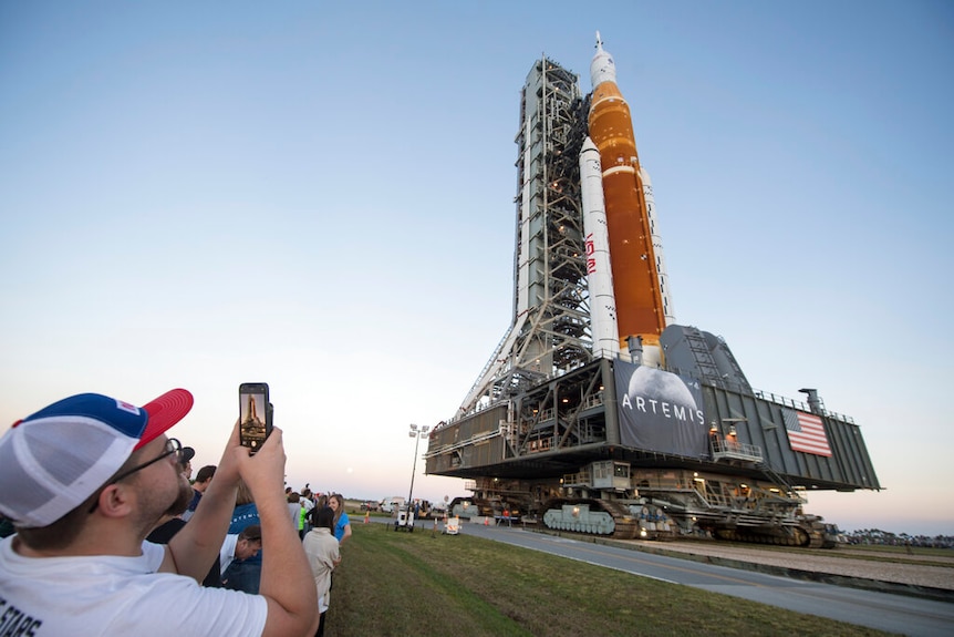 Un hombre toma una foto con su teléfono mientras un cohete del Sistema de Lanzamiento Espacial de la NASA y la nave espacial Orion a bordo ruedan por la carretera.