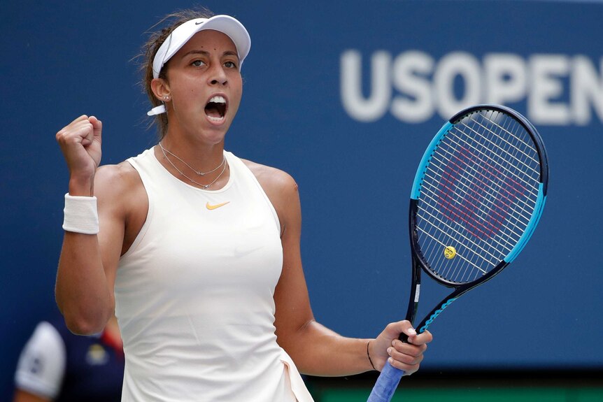 A female tennis player wearing a white top and visor clenches her fist and shouts.