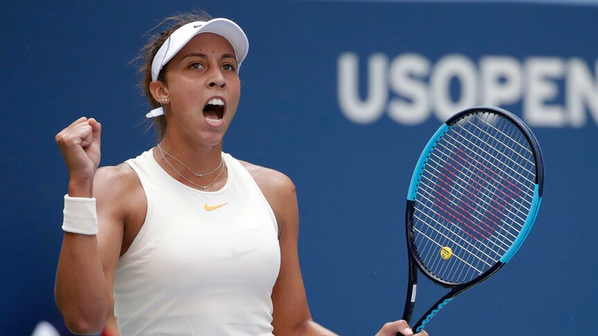 A female tennis player wearing a white top and visor clenches her fist and shouts.