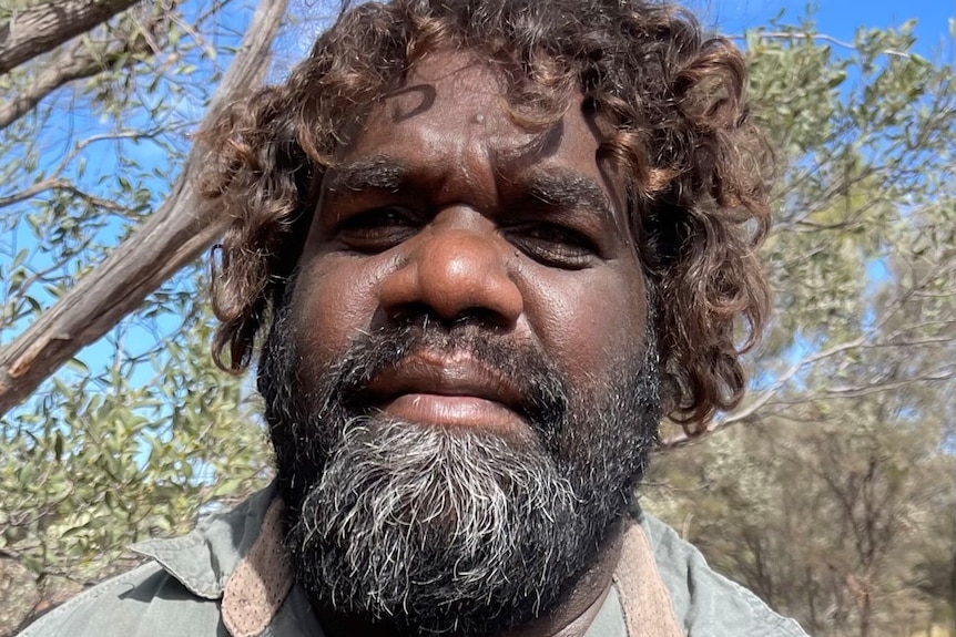 An Indigenous Australian man  with a beard and curly hair poses for a selfie.