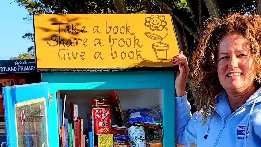 A woman stands next to brightly painted wooden box on a stand with books and cans of food