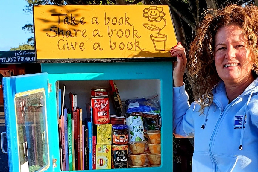 A woman stands next to brightly painted wooden box on a stand with books and cans of food