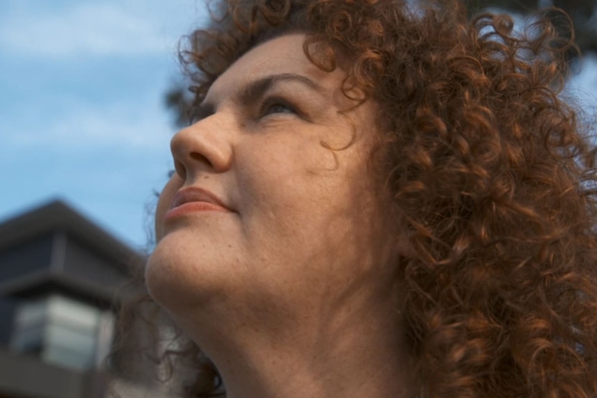 A close-up of a woman with brown curly hair, looking up at the sky.