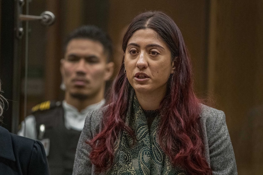 A woman wearing glasses and a headscarf looks of camera as she speaks in a court room.