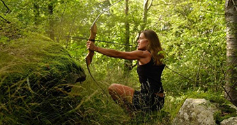 Side-profile of a woman squatting, holding up a bow and arrow, with a determined face, surrounded by forest.