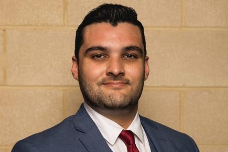 A mid shot of a young man wearing a grey suit, white shirt and red tie and smiling for a photo in front of a brick wall.