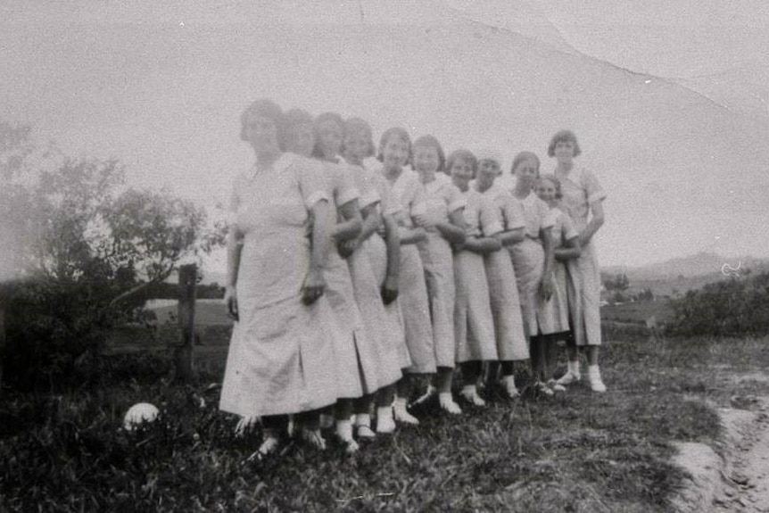 Black and white photo of 11 girls wearing school dresses standing in a line.