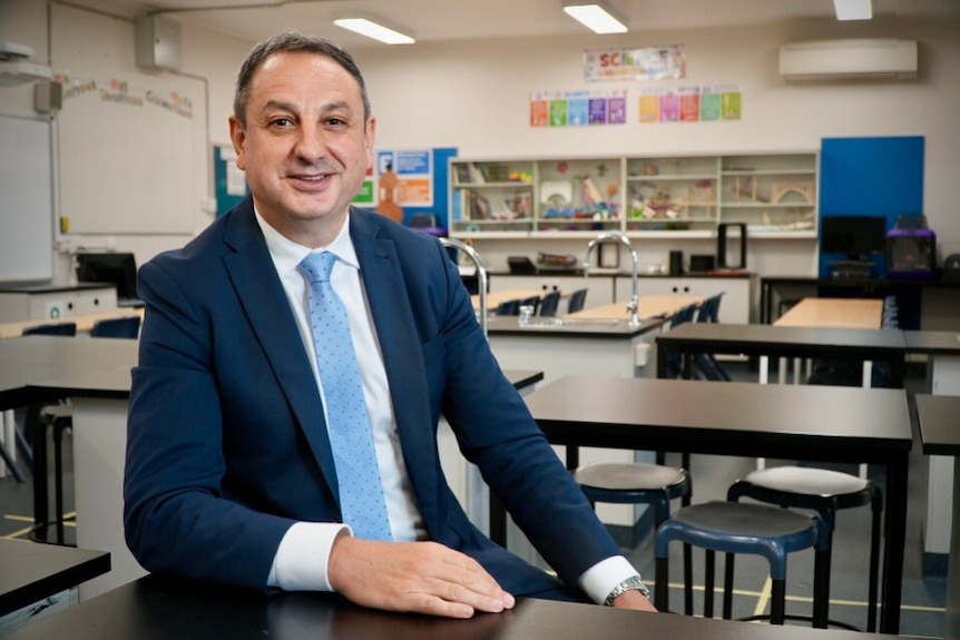 Man in navy suit and light blue tie smiles warmly in a school classroom.
