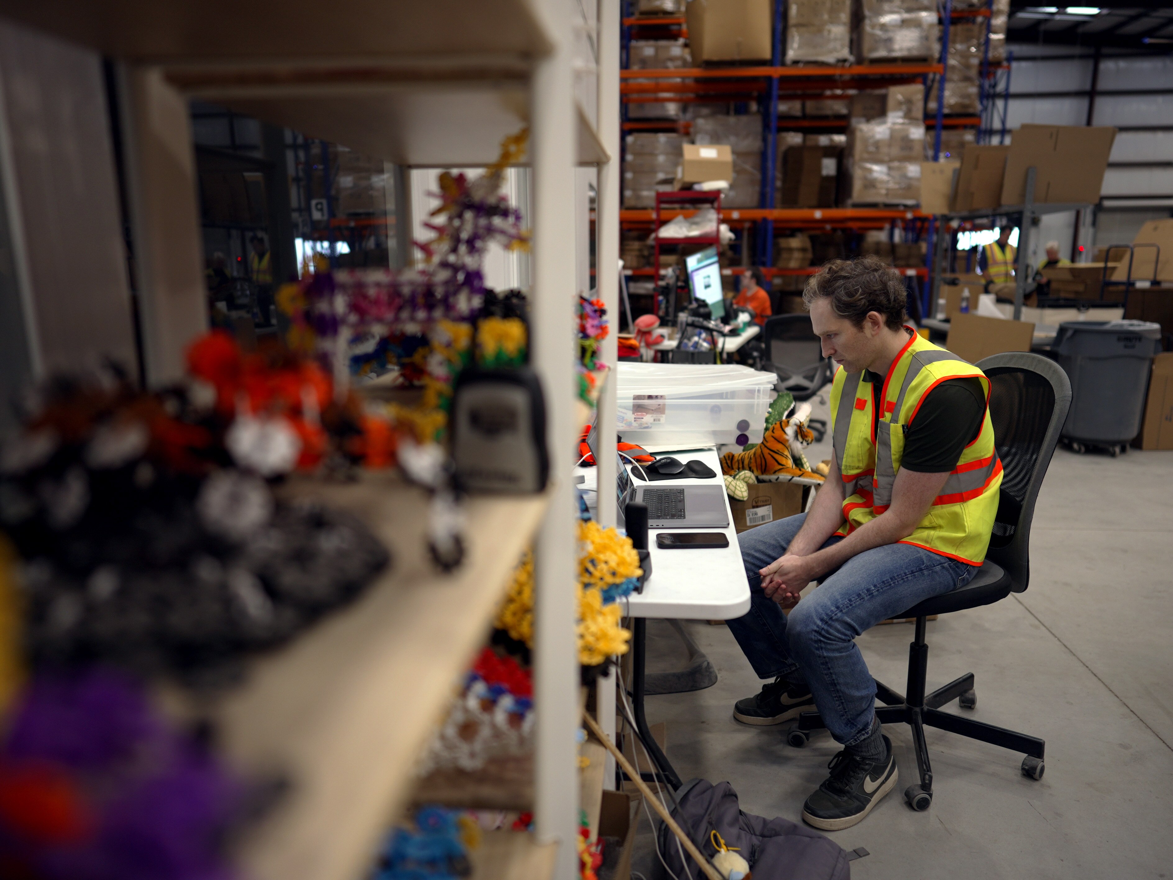 A man looks at a computer on a desk in a warehouse. On a shelf nearby are toys.