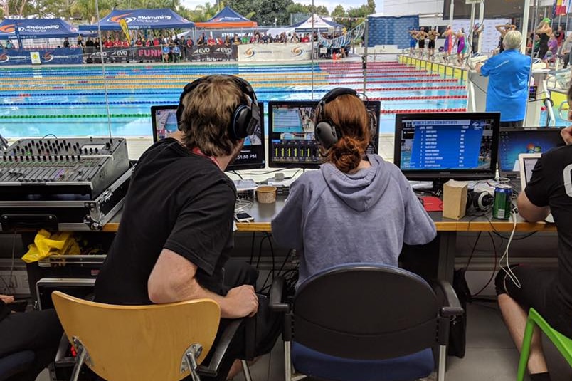 Three young people watch computers and, further ahead, a swimming pool where competitors are preparing to race.