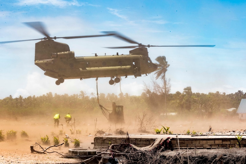 A Chinook helicopter flies low to the ground to drop off heavy equipment with dust everywhere and people ducking.