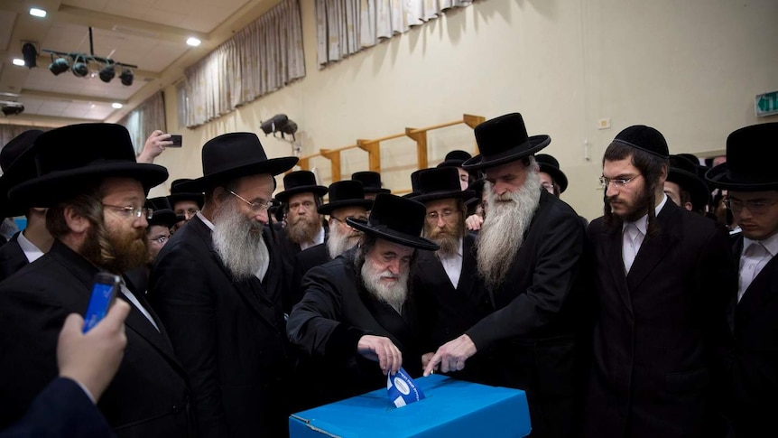 Male orthodox Jews clad in traditional black suits crowd around a blue voting box and one of them casts their vote.