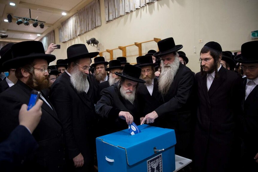 Male orthodox Jews clad in traditional black suits crowd around a blue voting box and one of them casts their vote.