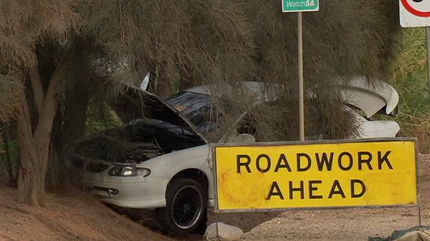 A white sedan crashed into a tree with a yellow roadwork ahead sign in front of it