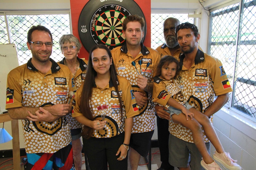 Six members of the Daley Darts team and a child stand in front of a darts board