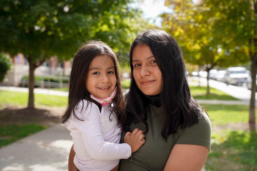 A woman with black hair and wearing a green shirt holds a little girl in a park.