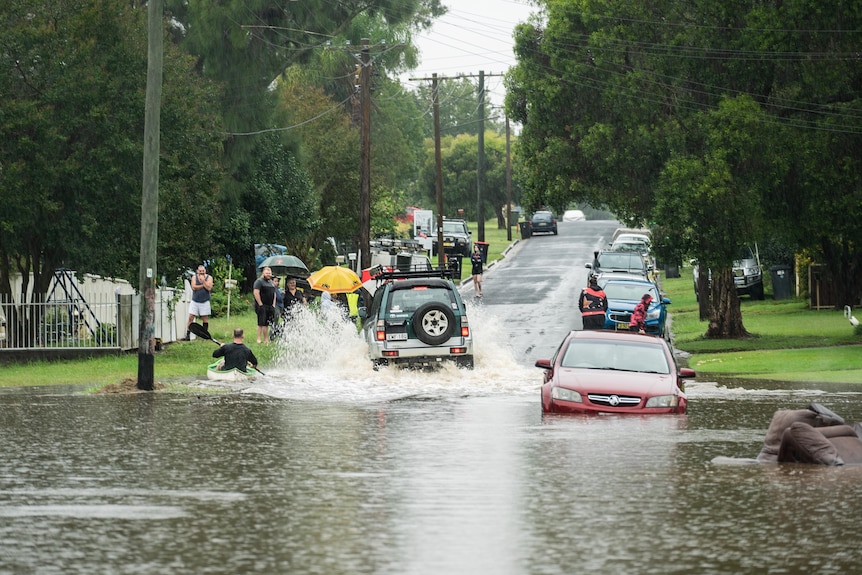 A car drives through a flooded street as people watch on.