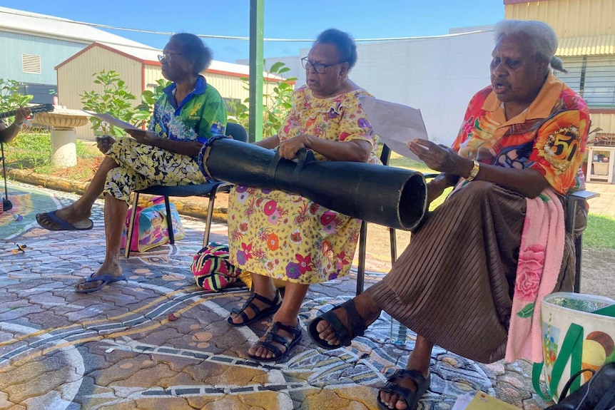 Three elderly women sit on chairs singing while one also plays a traditional hand drum