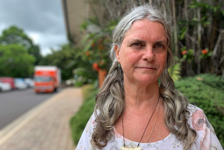 An older woman with long, grey hair stands on a street in a regional town.