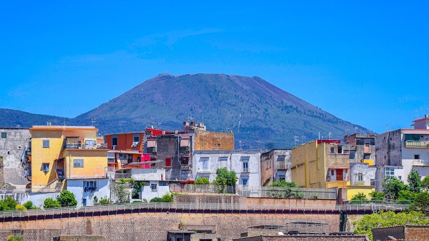 Small multi-coloured houses in the foreground with a grassy mount versuvius in the background with a blue sky above