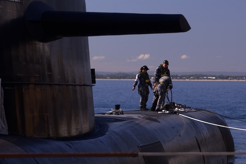 Three people wearing navy uniform stand on the deck of a large black submarine. which is sitting in water.