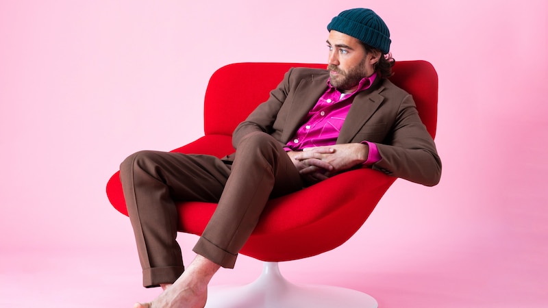 A 2022 press shot of Matt Corby - barefoot in a suit and beanie - relaxed in a red lounge chair on a pink background