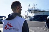 In the foreground we see the back of a man, wearing a bib with the MSF logo. He looks towards a big ship docked