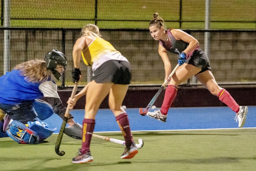 Angie Lambert holds a hockey stick and jumps in the air as two other players fight for the puc. 