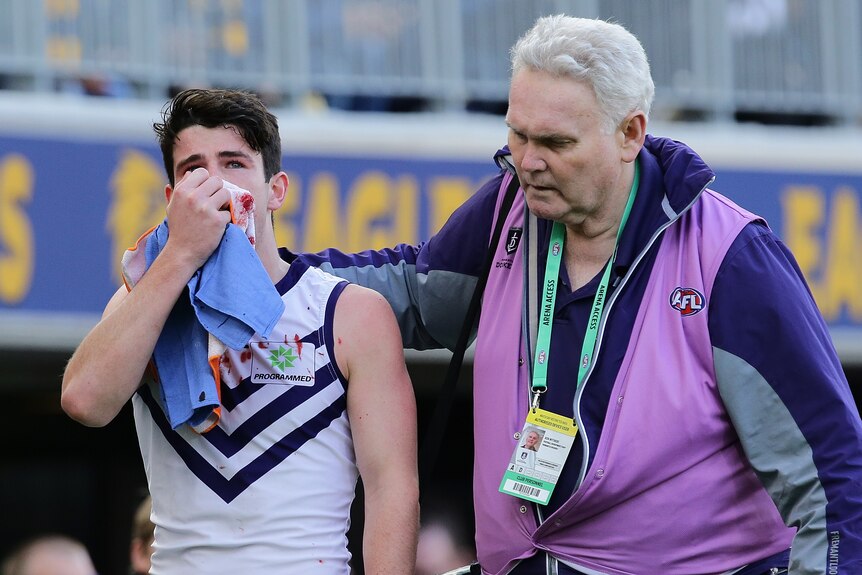 A Fremantle AFL player walks off with a blood-stained towel held to his face, as a trainer puts his arm around him. 