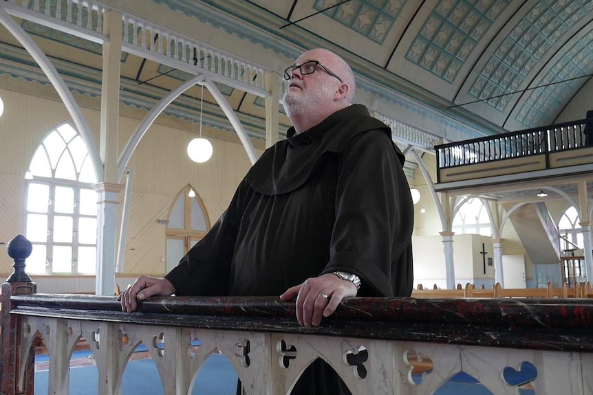 A priest leans on a wooden railing and stares at the ceiling of a timber church