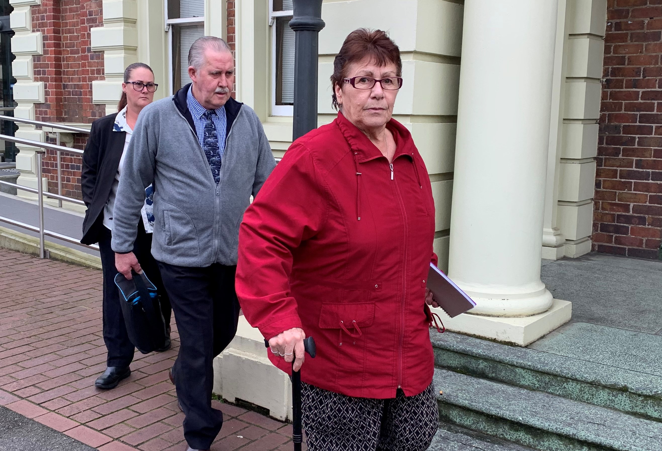 A woman in a red sweater looks at the camera as she leaves a court building followed by a man and another woman